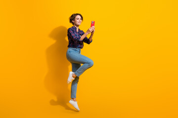 Fototapeta na wymiar Full size photo of charming girl wear checkered blouse jumping enjoy fast internet speed on smartphone isolated on yellow color background