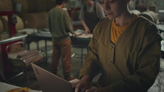 Tilting up shot of young Caucasian female supervisor using laptop placed on stack of freshly produced paper while other female employees working at production line in printing houseTilting up shot of 