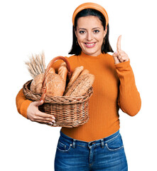 Young hispanic girl holding wicker basket with bread smiling with an idea or question pointing...