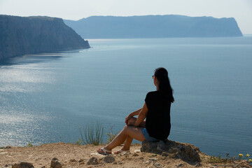 A beautiful woman with dark hair sits on the edge of a mountain against the backdrop of the sea and sky. View from the back