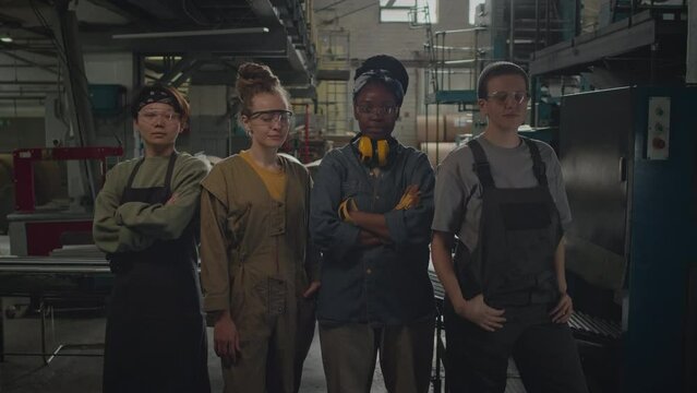 Zoom-in medium lowkey portrait of ethnically diverse team of four young female factory workers standing confidently between machine equipment and looking at cameraZoom-in medium lowkey portrait of eth