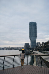 A young woman admires the views in the modern Waterfront area, near the Sava River embankment. View from the ground in a snowless gray winter. Rear view. Belgrade, Serbia - 07.01.2024.