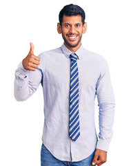 Young latin man wearing business clothes smiling happy and positive, thumb up doing excellent and approval sign