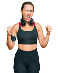 Beautiful blonde woman wearing gym clothes and using headphones angry and mad raising fists frustrated and furious while shouting with anger. rage and aggressive concept.