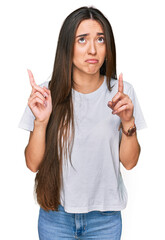 Young hispanic girl wearing casual white t shirt pointing up looking sad and upset, indicating...
