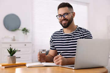 Young man with glass of water writing in notebook at wooden table indoors