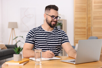 Fototapeta premium Young man writing in notebook while working on laptop at wooden table indoors
