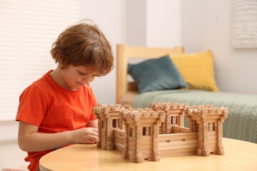 Cute little boy playing with wooden fortress at table in room. Child's toy