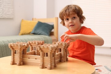 Cute little boy playing with wooden fortress at table in room. Child's toy