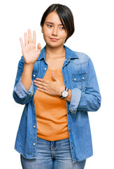 Young beautiful hispanic woman with short hair wearing casual denim jacket swearing with hand on...