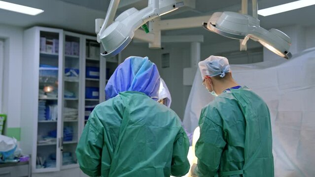 Rear view of two surgeons wearing uniforms standing near the operational table. Doctors carry out the trauma surgery on a leg.