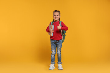 Happy schoolgirl with backpack and books showing thumb up gesture on orange background