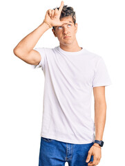 Fototapeta premium Young handsome man wearing casual white tshirt making fun of people with fingers on forehead doing loser gesture mocking and insulting.