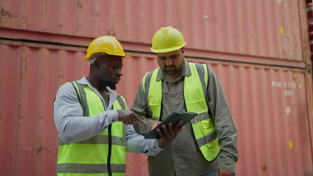 Two industry worker in safety uniform checking container on tablet together at warehouse. Black male supervisor and Indian industrial engineer talking, working on tablet. Shipping industry, Logistics