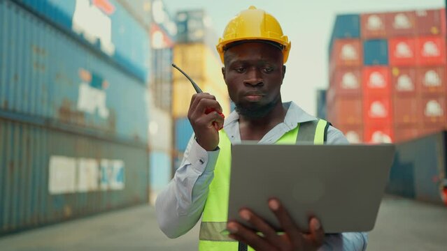 Black African foreman industrial engineer in hard hat and safety vest working on laptop and radio phone calling team worker in containers warehouse. Shipping import export industry Logistics business
