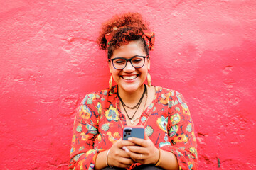 Fototapeta premium A vibrant portrait of a joyful young woman smiling while texting on her mobile phone against a vivid red background.