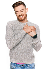 Young redhead man wearing casual winter sweater smiling with hands on chest with closed eyes and grateful gesture on face. health concept.