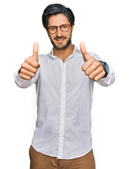 Young hispanic man wearing business shirt and glasses approving doing positive gesture with hand, thumbs up smiling and happy for success. winner gesture.