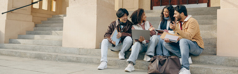 Happy students sitting on stairs, looking at laptop and discussing their project together