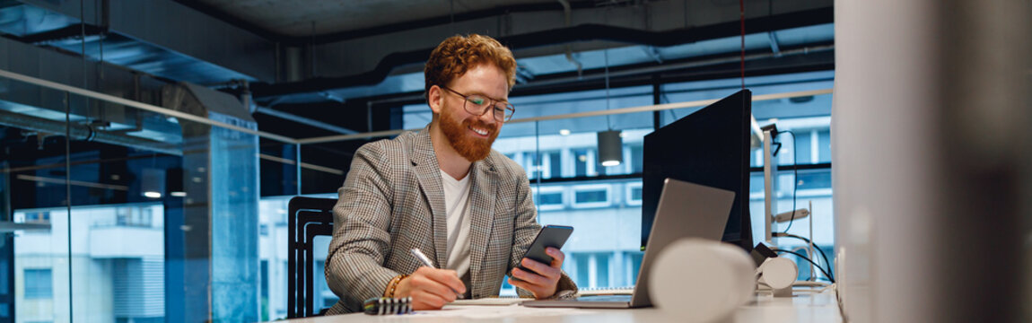 Smiling male businessman looking on phone while working on laptop in modern office space