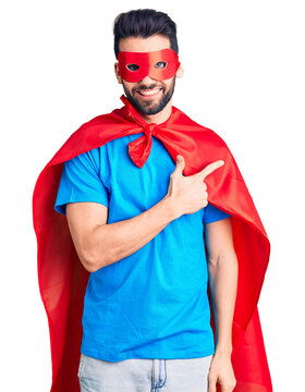 Young handsome man with beard wearing super hero costume cheerful with a smile of face pointing with hand and finger up to the side with happy and natural expression on face