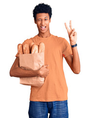 Young african american man holding paper bag with bread smiling with happy face winking at the...