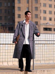 Portrait of handsome Chinese young man with black short hair wearing woolen coat posing with modern city building background in sunny winter day, male fashion, cool Asian young man.