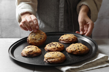 Woman taking delicious chocolate chip cookie from tray at table, closeup