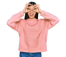 Beautiful young asian woman wearing casual winter sweater doing ok gesture like binoculars sticking tongue out, eyes looking through fingers. crazy expression.