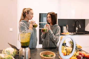 mother with her teenage daughter drinking healthy green smoothie recording vlog video and live...