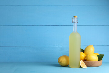 Bottle of tasty limoncello liqueur, lemons and green leaves on light blue table, space for text