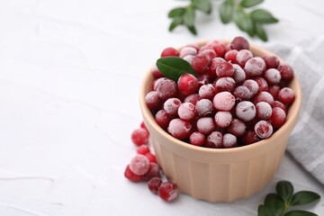Frozen red cranberries in bowl and green leaves on white table, closeup. Space for text