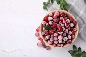 Frozen red cranberries in bowl and green leaves on white table, top view. Space for text