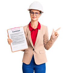 Young beautiful woman wearing architect hardhat holding clipboard with contract document smiling happy pointing with hand and finger to the side