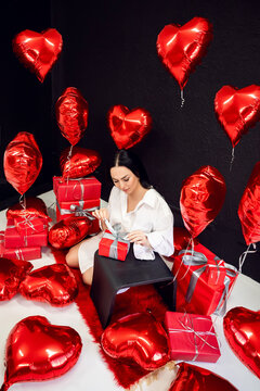 brunette woman in a white shirt on the background of balloons of hearts unpacks gifts. The concept of Birthday and Valentine's Day.