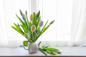 a bouquet of tulips in a ceramic teapot with decorative colored eggs on the window sill. Home decor for Easter