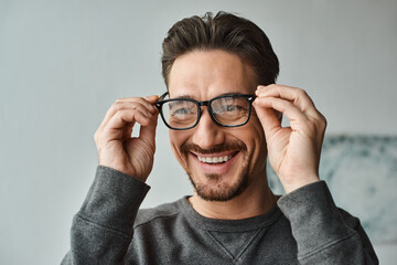 portrait of cheerful and bearded man in grey jumper wearing eyeglasses and looking at camera