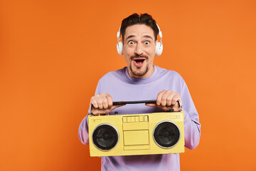 surprised bearded man in purple sweater and wireless headphones holding boombox on orange background