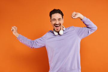 Naklejka premium joyful man in purple sweater with wireless headphones posing on orange background, music lover