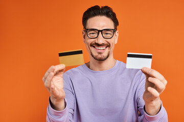 cheerful bearded man in glasses holding credit cards on orange background, shopping and consumerism