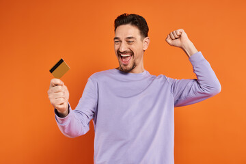 excited man with beard smiling and holding credit card on orange background, rejoicing gesture