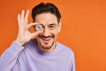 happy man holding bitcoin over his eye and smiling at camera on orange background, cryptocurrency