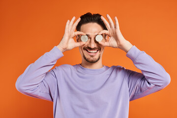 happy man holding bitcoins over his eyes and smiling at camera on orange background, cryptocurrency