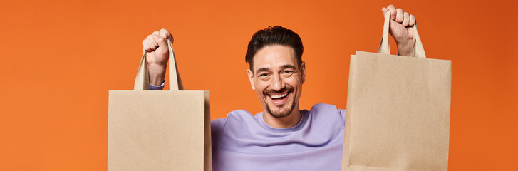 happy bearded man in casual attire holding shopping bags on orange background, consumerism banner