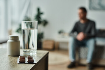 medication in bottle and blister pack near glass of water on table and blurred man on background - obrazy, fototapety, plakaty