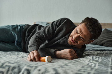 bearded man suffering from pain and lying on bed near bottle with medication, cramp and discomfort