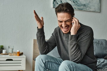 angry bearded man in grey sweater talking on smartphone and gesturing in modern bedroom, argue