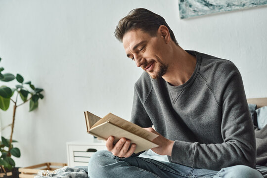 pleased man in grey casual jumper reading book while relaxing on weekend in bedroom, leisure