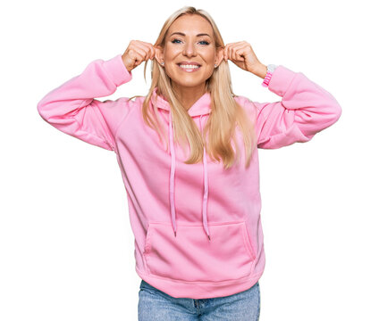 Young blonde woman wearing casual sweatshirt smiling pulling ears with fingers, funny gesture. audition problem