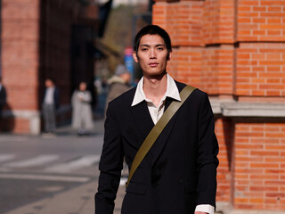 Portrait of handsome Chinese young man with black short hair wearing black blazer posing with modern city building background in sunny winter day, male fashion, cool Asian young man.